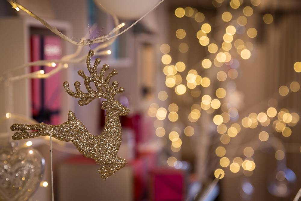 A close up image of a reindeer Christmas decoration hanging on a tree, with blurry lights in the background